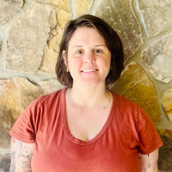 Woman with short brown hair smiles, wearing an orange v-neck shirt, in front of a stone wall.