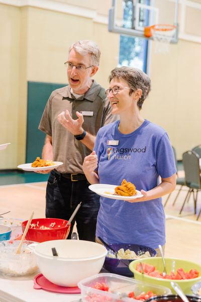 Two people holding plates of food at a table with other dishes, possibly a church event in a gym.