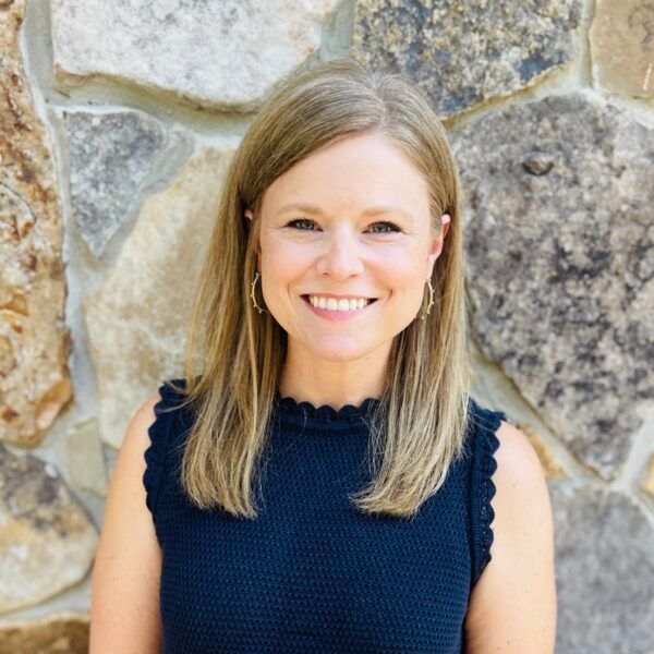 Woman smiling, posing in front of a stone wall. She wears a navy sleeveless top and small hoop earrings.