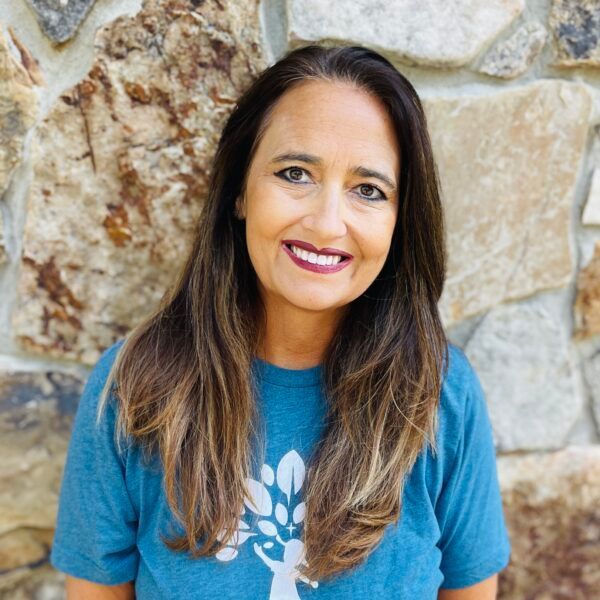 Woman with brown hair smiling in front of a stone wall, wearing a blue shirt with a white graphic.