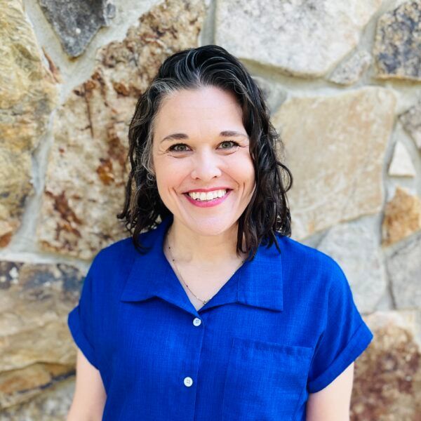 Woman with dark hair smiles, wearing a blue shirt, in front of a stone wall.
