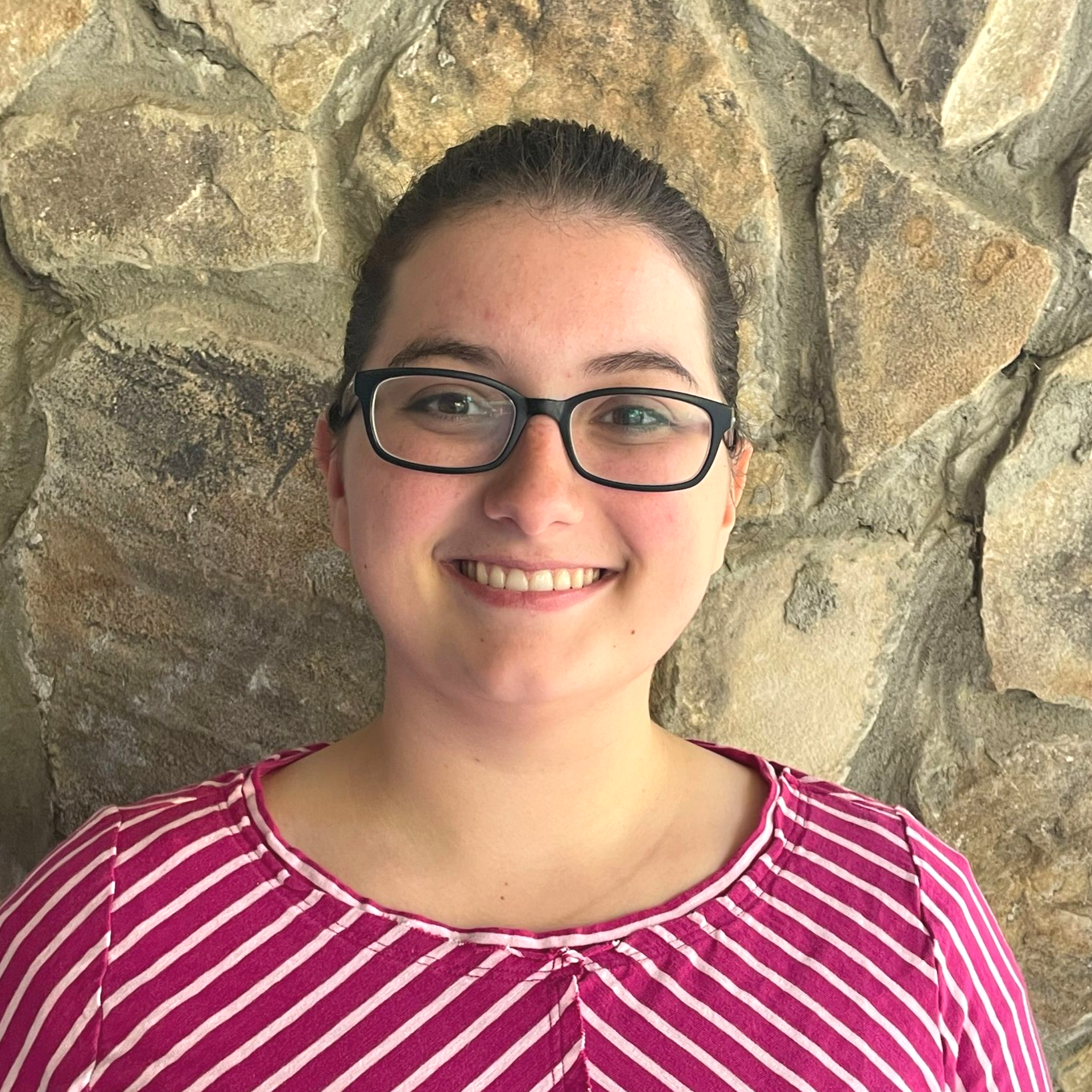 Woman with glasses smiles in front of a stone wall, wearing a pink striped shirt.