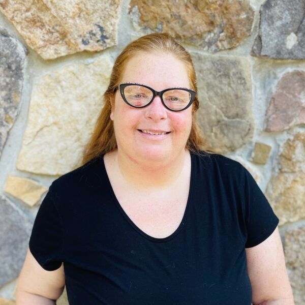 Woman with glasses smiles in front of a stone wall, wearing a black top.