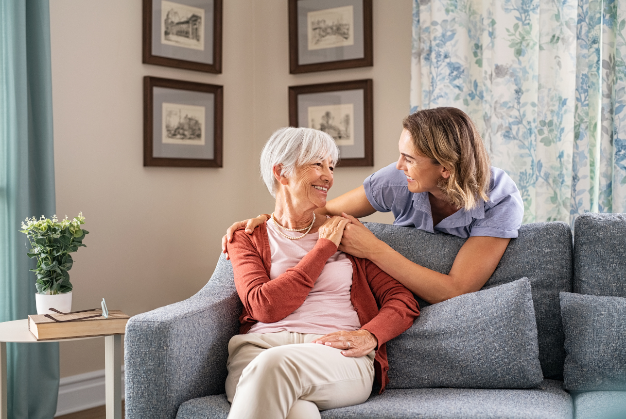 Woman on couch with caregiver, smiling. Living room setting, art on wall.