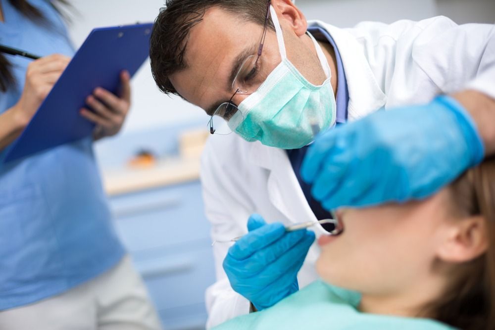 Dentist examining patient's teeth with mask and gloves; assistant takes notes in the office.