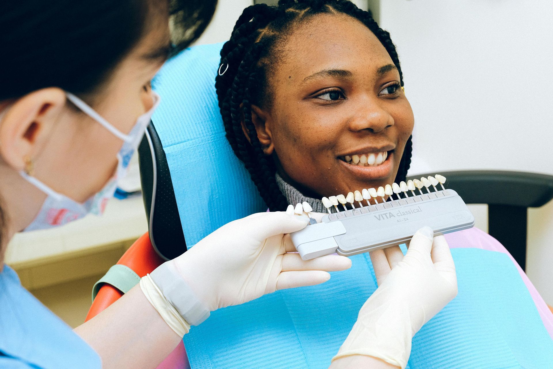 Dentist holding shade guide up to patient's teeth in dental chair; office setting.