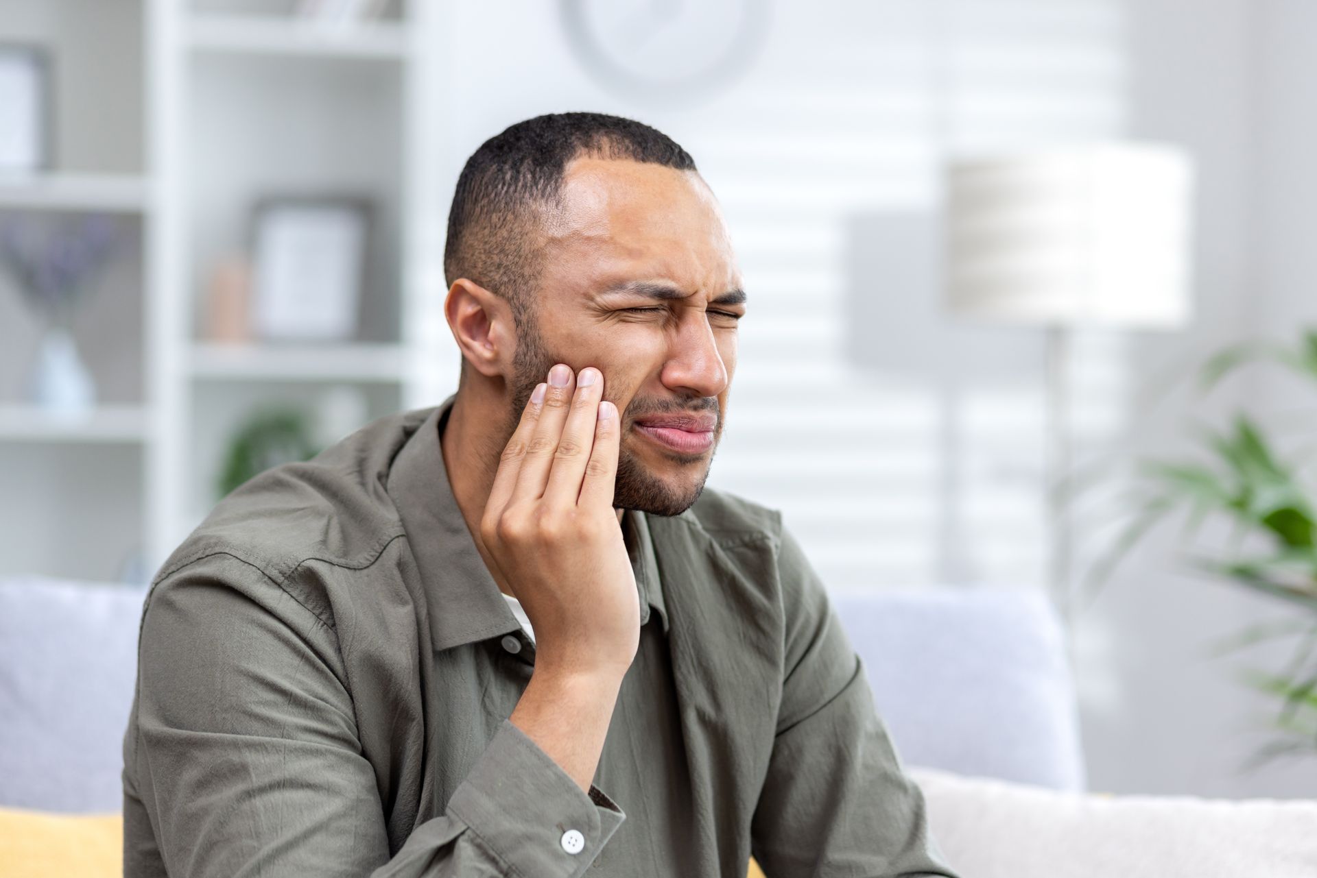 Man holding his cheek in discomfort while sitting on a couch in a bright living room