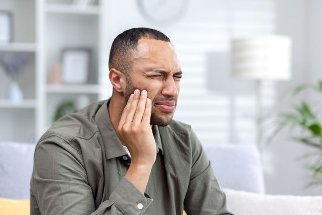 Person sitting on a sofa, touching their cheek with a pained expression in a bright living room.