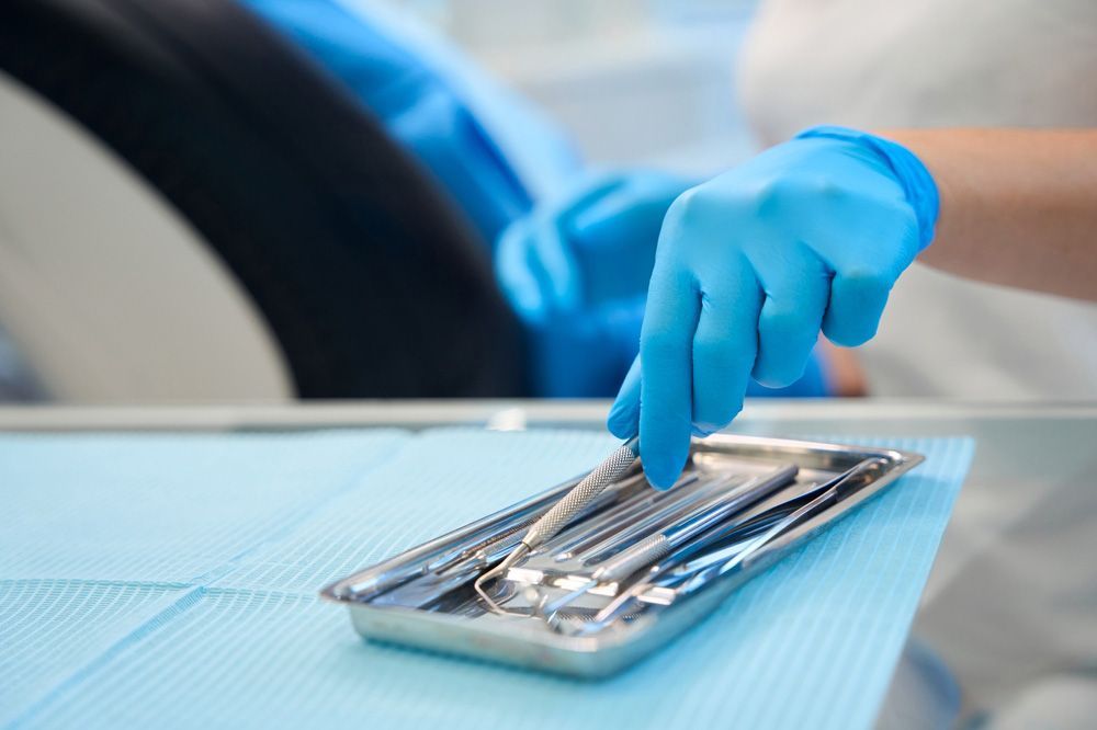 Dentist's gloved hand selecting a dental tool from a tray on a light blue surface.