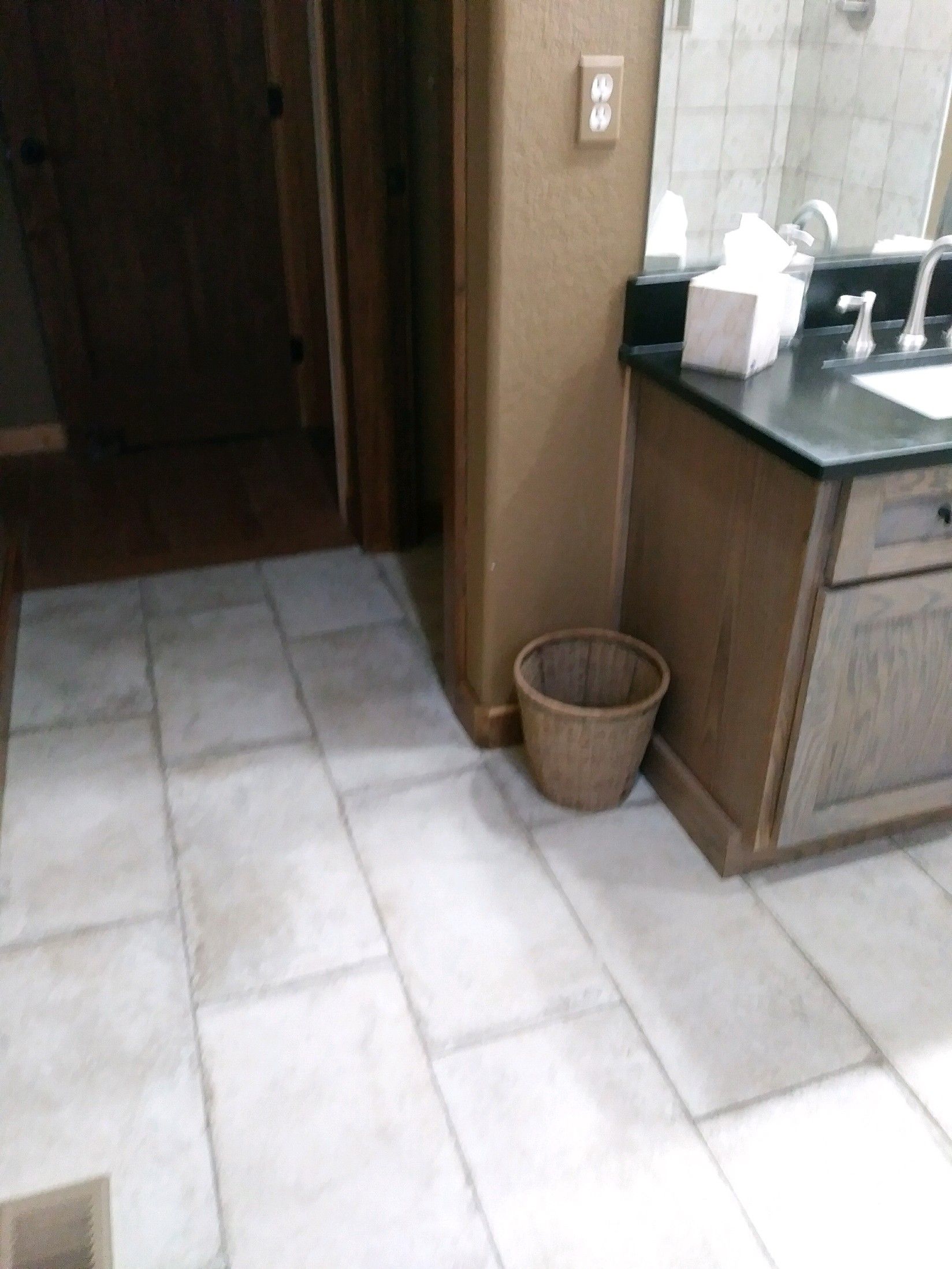 Bathroom with tiled floor, wooden vanity, and woven trash can. Doorway visible in the background.