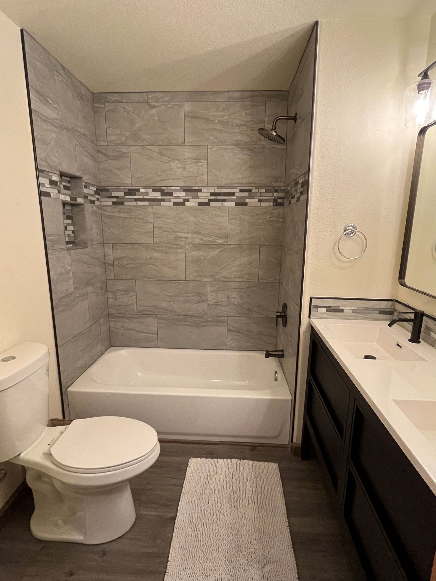 Bathroom with a gray tiled shower, white tub, dark vanity, and toilet.