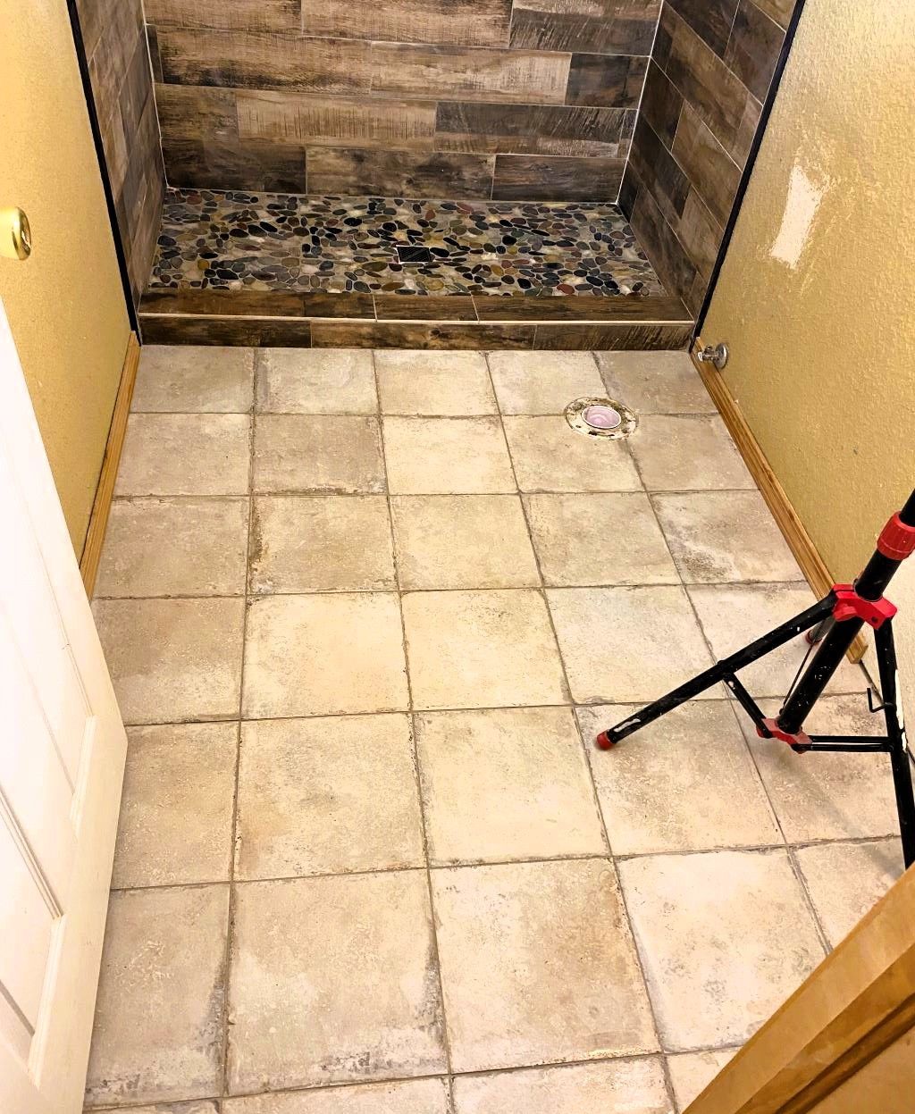 Bathroom floor with light-colored square tiles. Shower with dark stone floor and wood-look tiled walls is in the background.