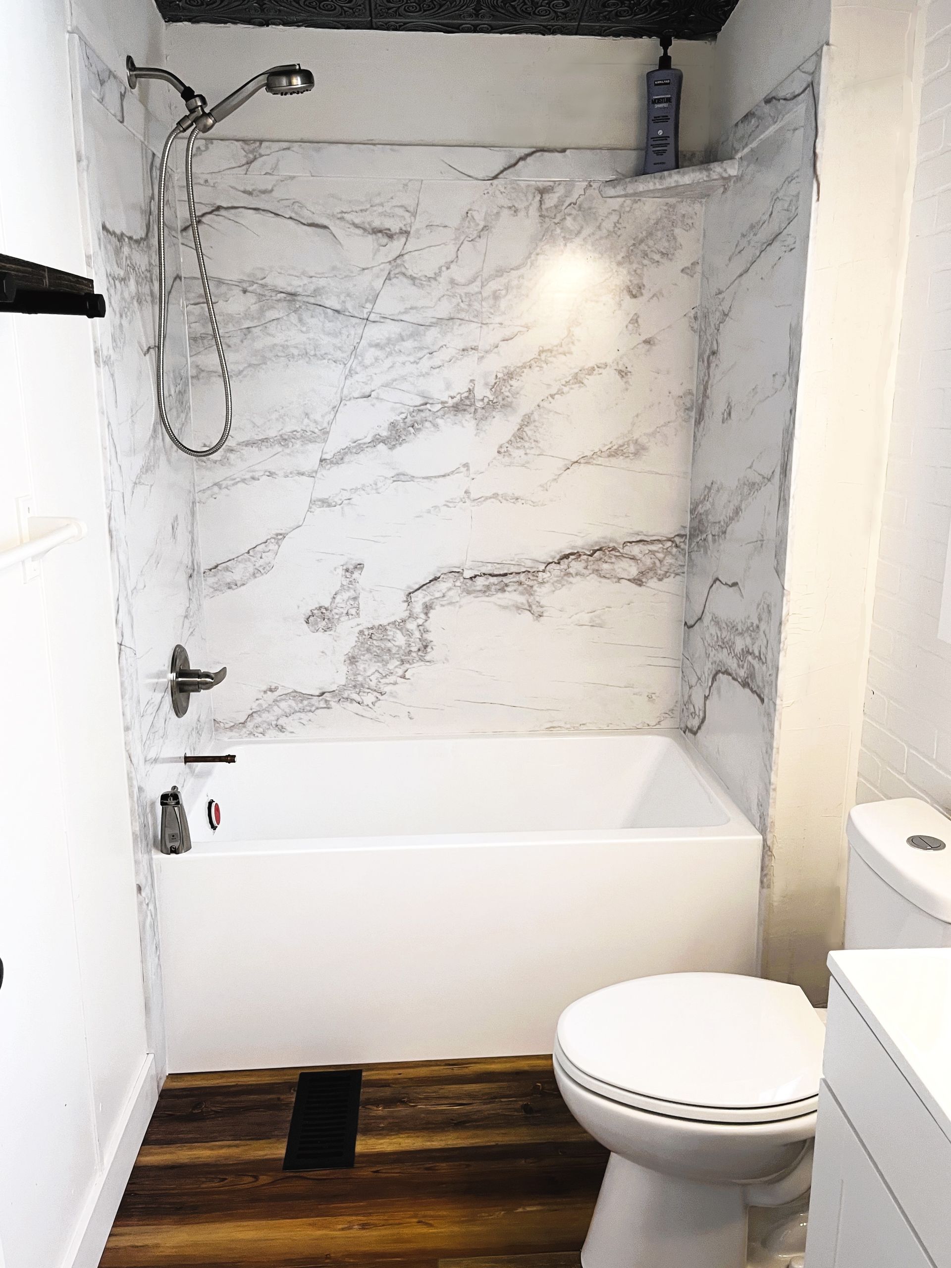 Small bathroom with a white tub, marble-patterned shower wall, and wooden floor. Toilet is beside the tub.