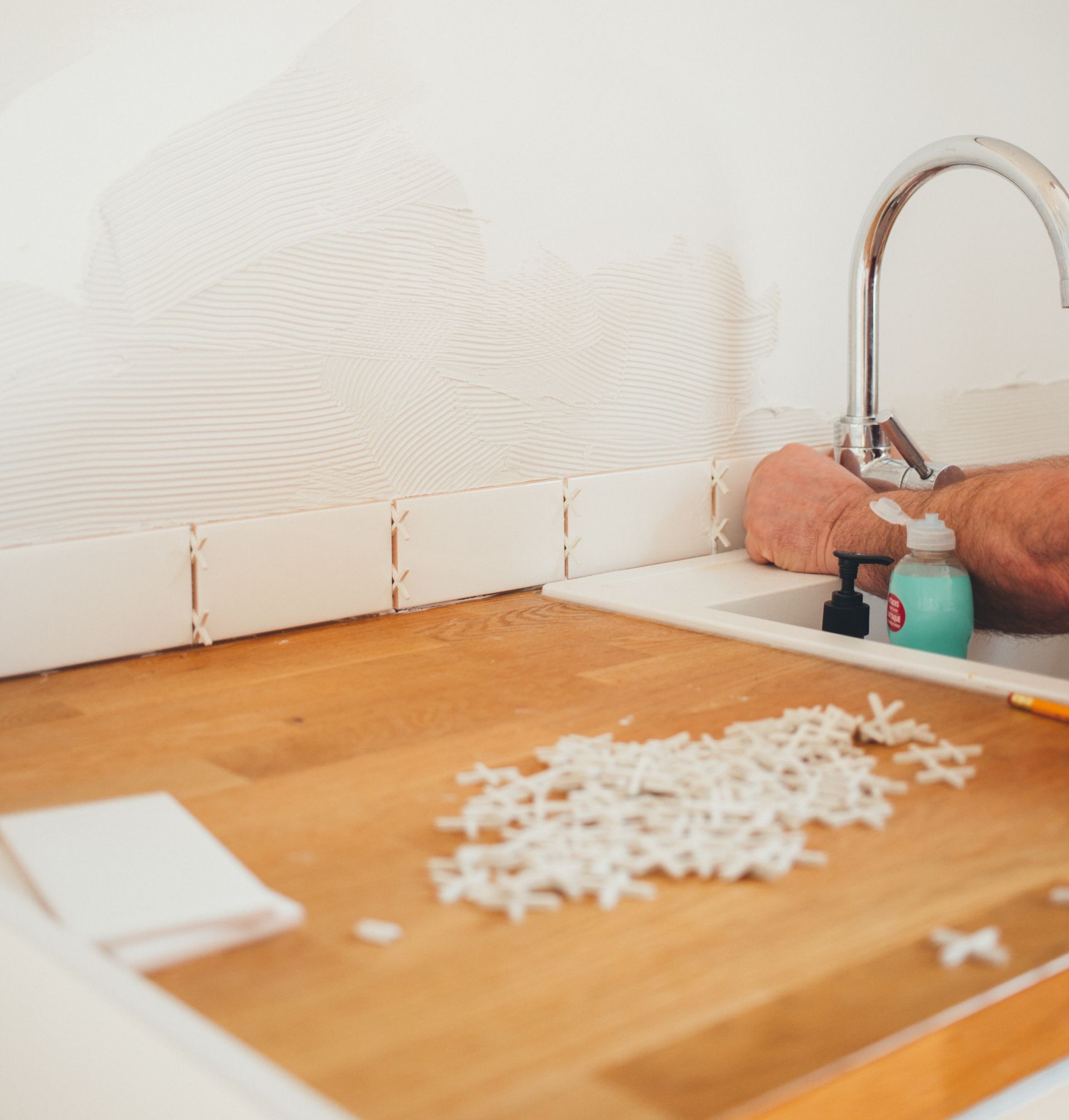 Kitchen counter with tiling in progress; white tiles, wooden counter, sink, and tile spacers.