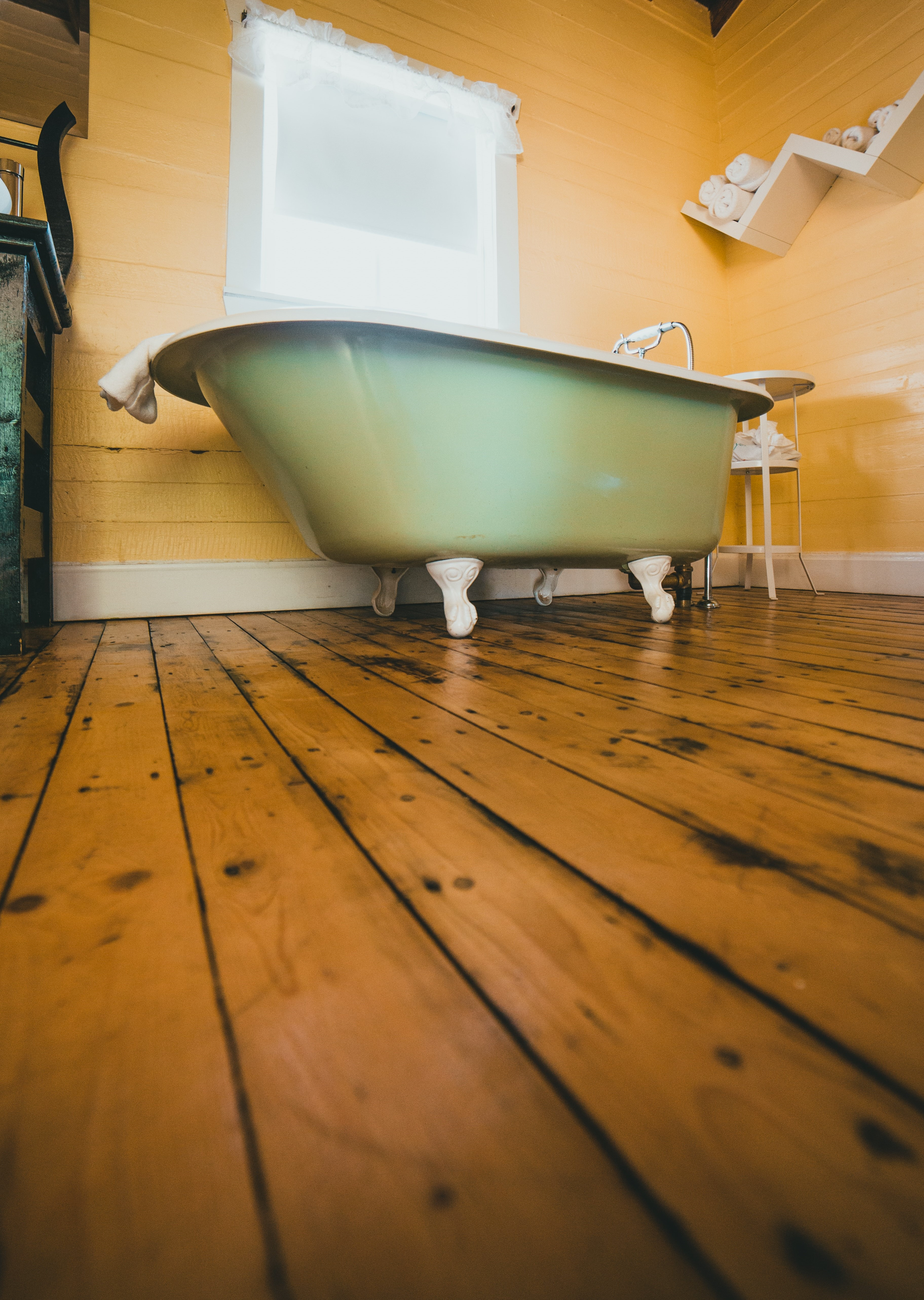 Light green claw-foot bathtub in a room with wood floors and yellow walls. A window is behind the tub.
