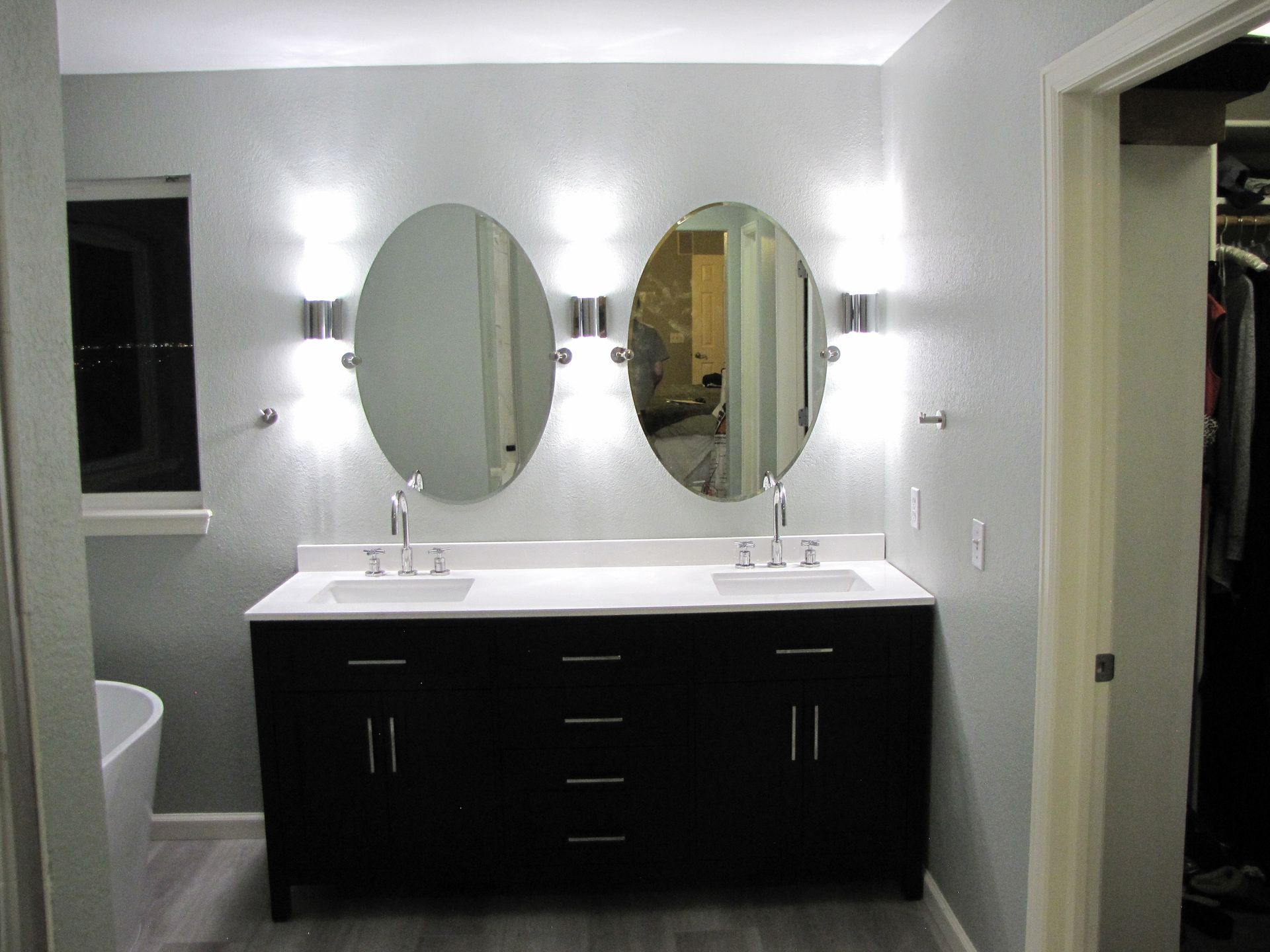Bathroom with a dark vanity, oval mirrors, and sconce lighting. Gray walls and flooring.