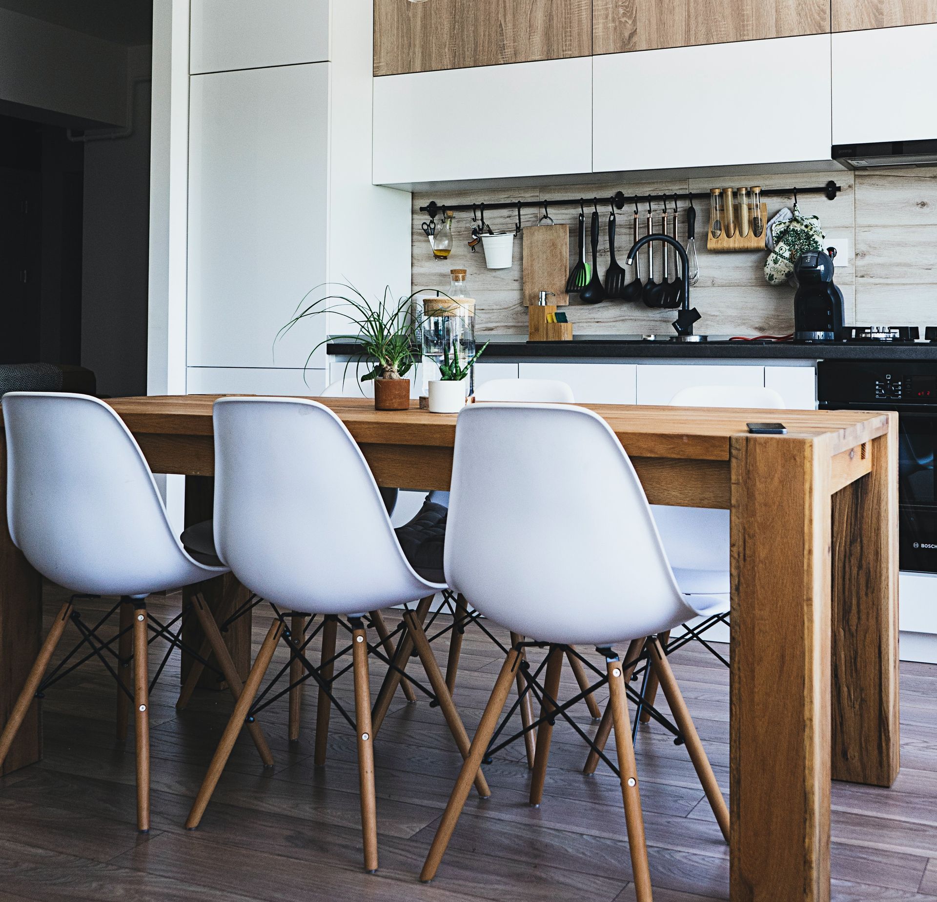 Dining area with a wooden table and three white chairs in front of a modern kitchen.