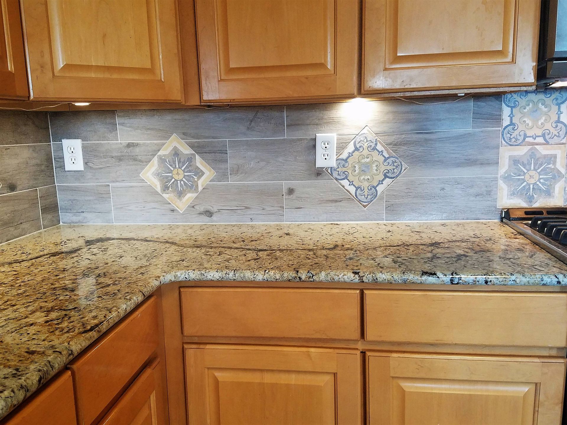 Kitchen backsplash with grey wood-look tiles and decorative patterned tiles, beige cabinets, and granite countertops.