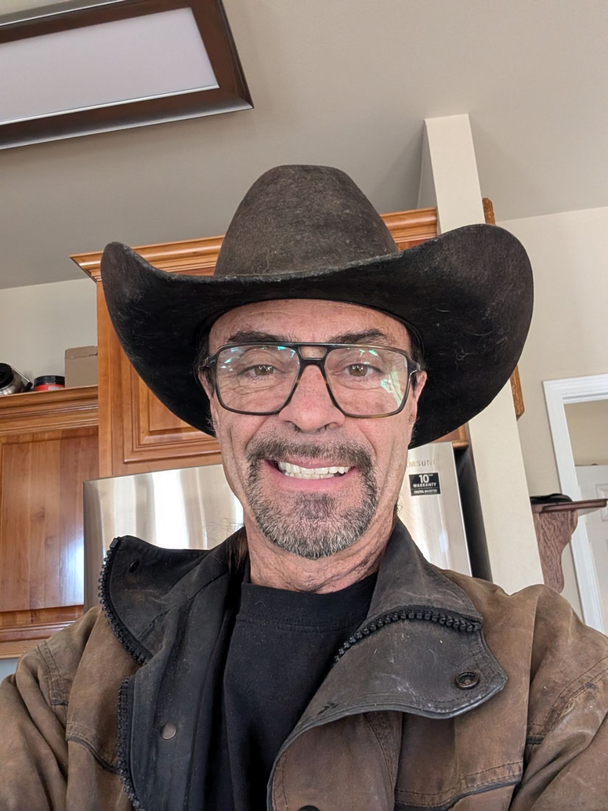 Man wearing cowboy hat and glasses, smiling, in front of a kitchen.
