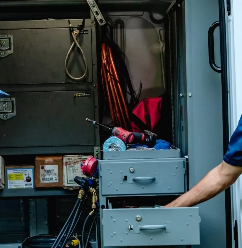 A person's hand reaching into a gray drawer in a work truck, tools and equipment visible in the cabinet.