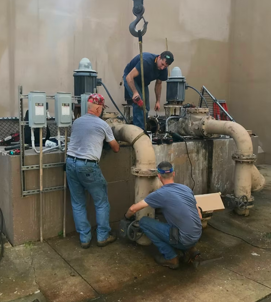 Three workers disassemble machinery in an industrial setting. Beige walls, metal pipes, and overhead crane.