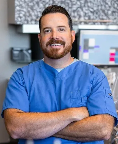 Man in blue scrubs with crossed arms, smiling, in a medical setting.