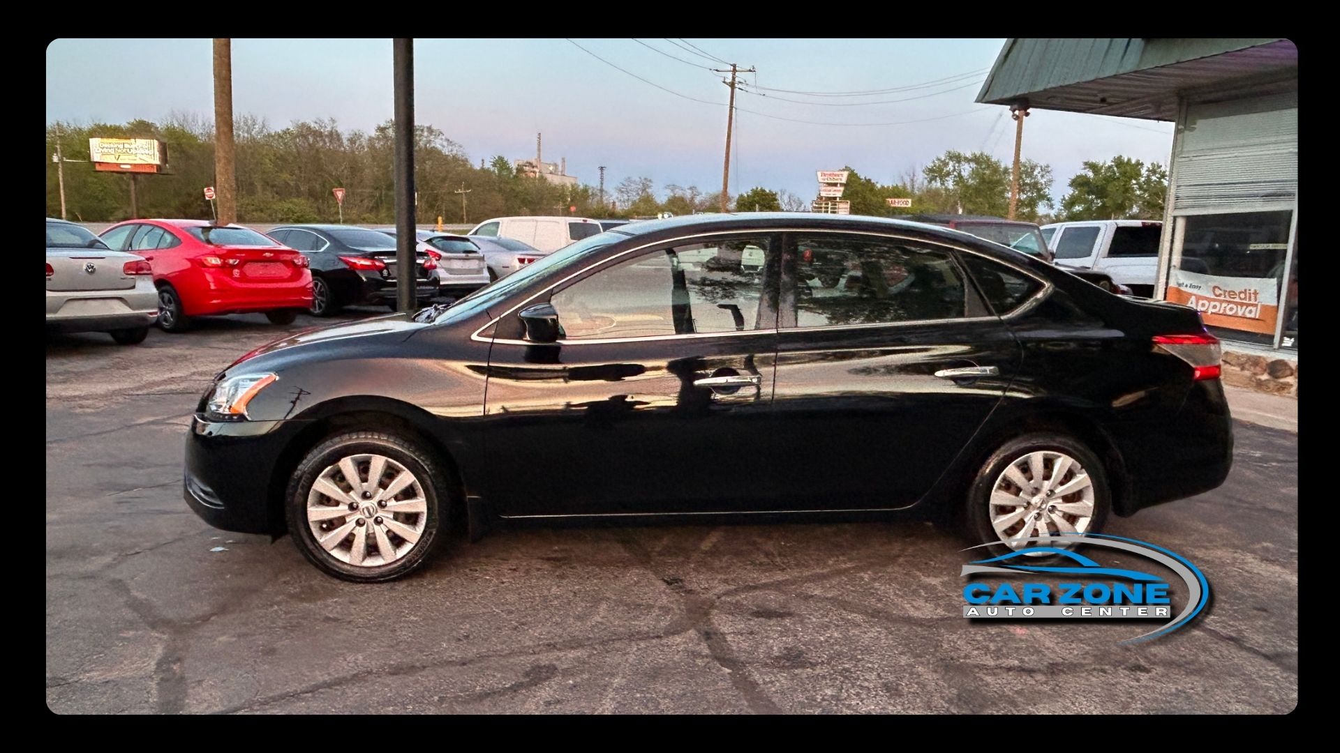 Black sedan parked on a car lot, side view, with other vehicles and dealership signage in the background.