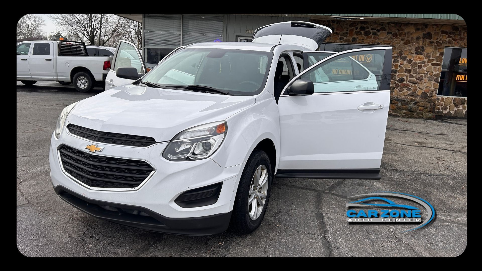 White Chevrolet SUV parked with driver door open in a lot, rear hatch open and a truck in the background