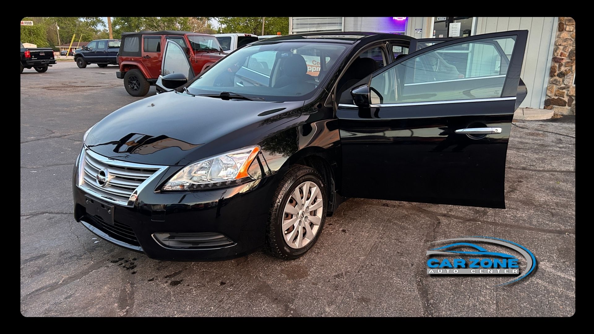 Black sedan with front passenger door open, parked outdoors on gravel near other vehicles.