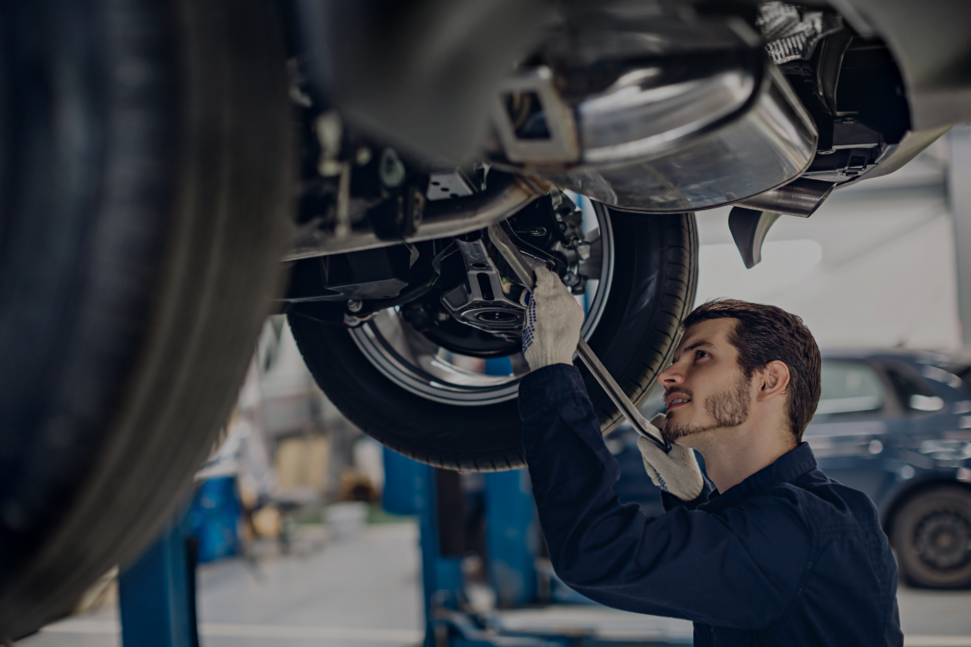 Mechanic working under a car in a garage, wearing gloves and a navy jumpsuit. at Car Zone Auto Center 2344 Sagamore Pkwy N, Lafayette IN 47905