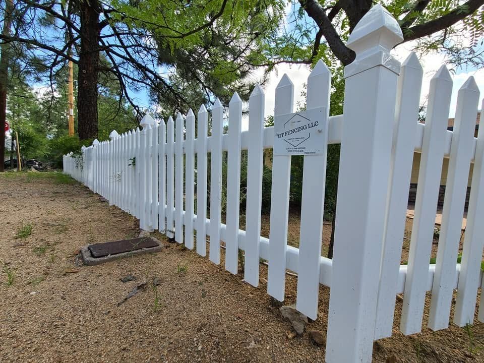 White picket fence along a dirt path beside trees and a small sign outdoors.