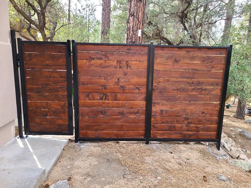 Wooden backyard fence with black metal frame and gate beside a house, set among trees and gravel ground