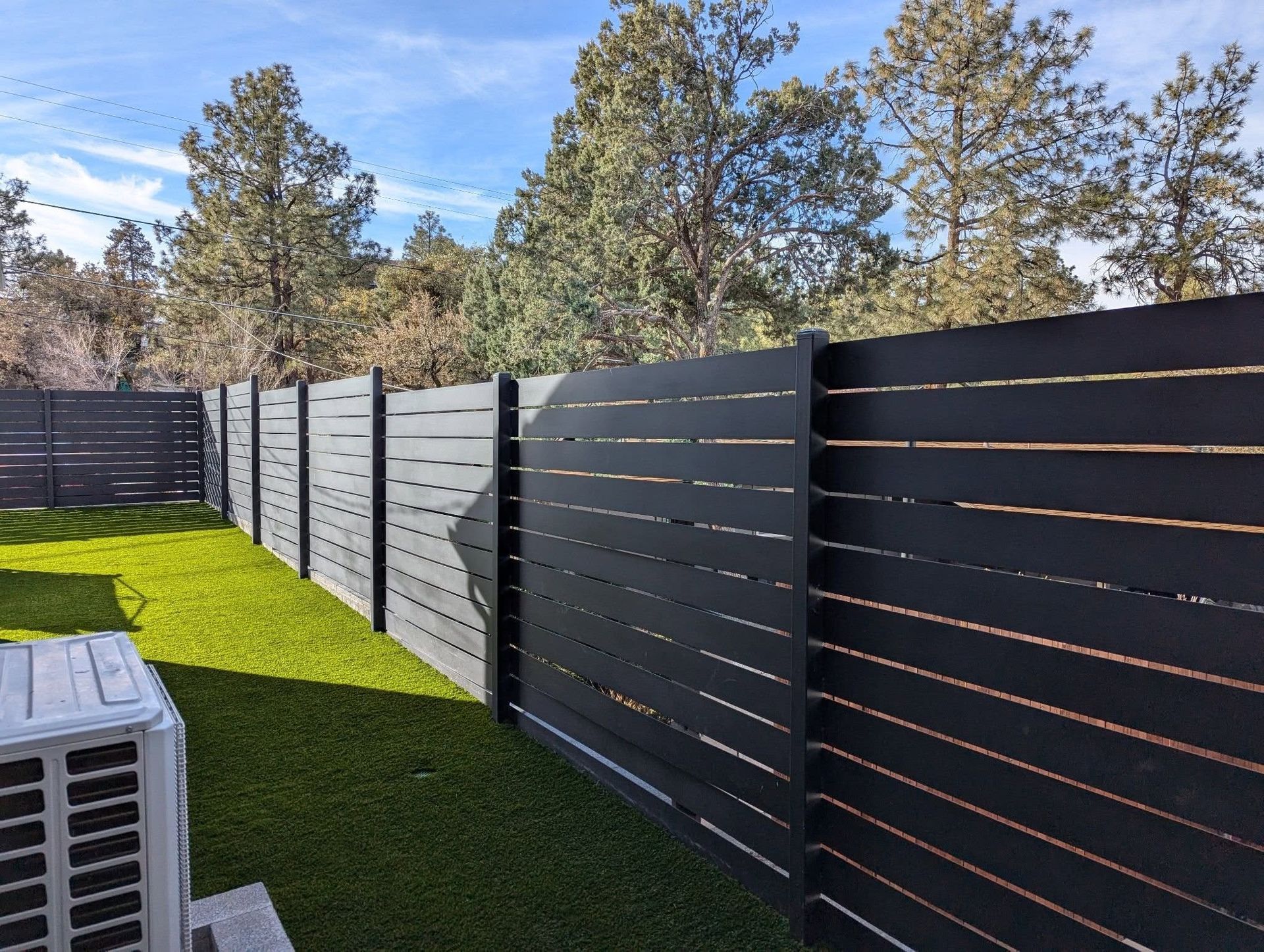 Black slat fence along a green lawn with pine trees and blue sky in the background