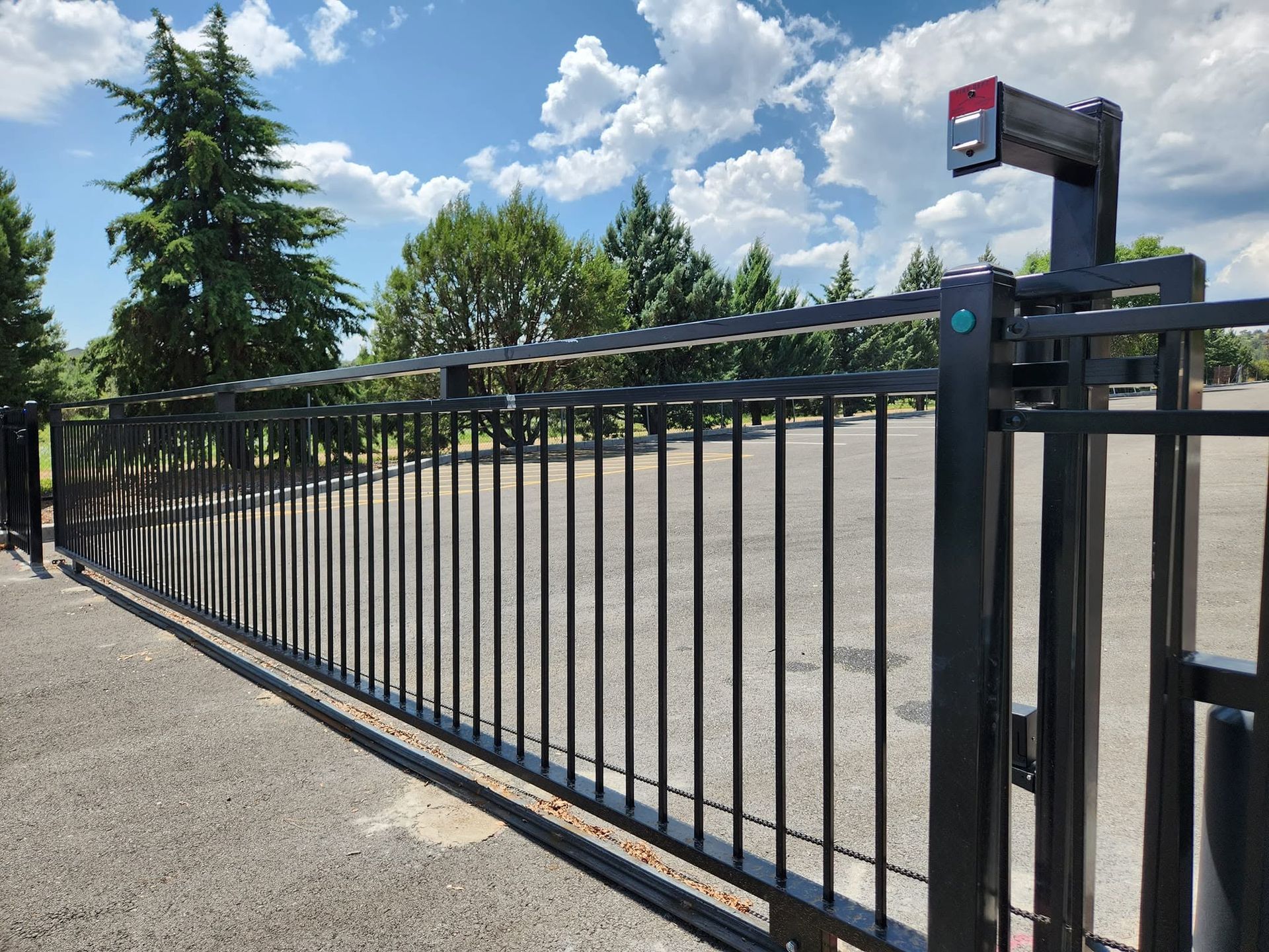 Black metal parking gate beside a paved lot, with trees and a blue sky in the background.
