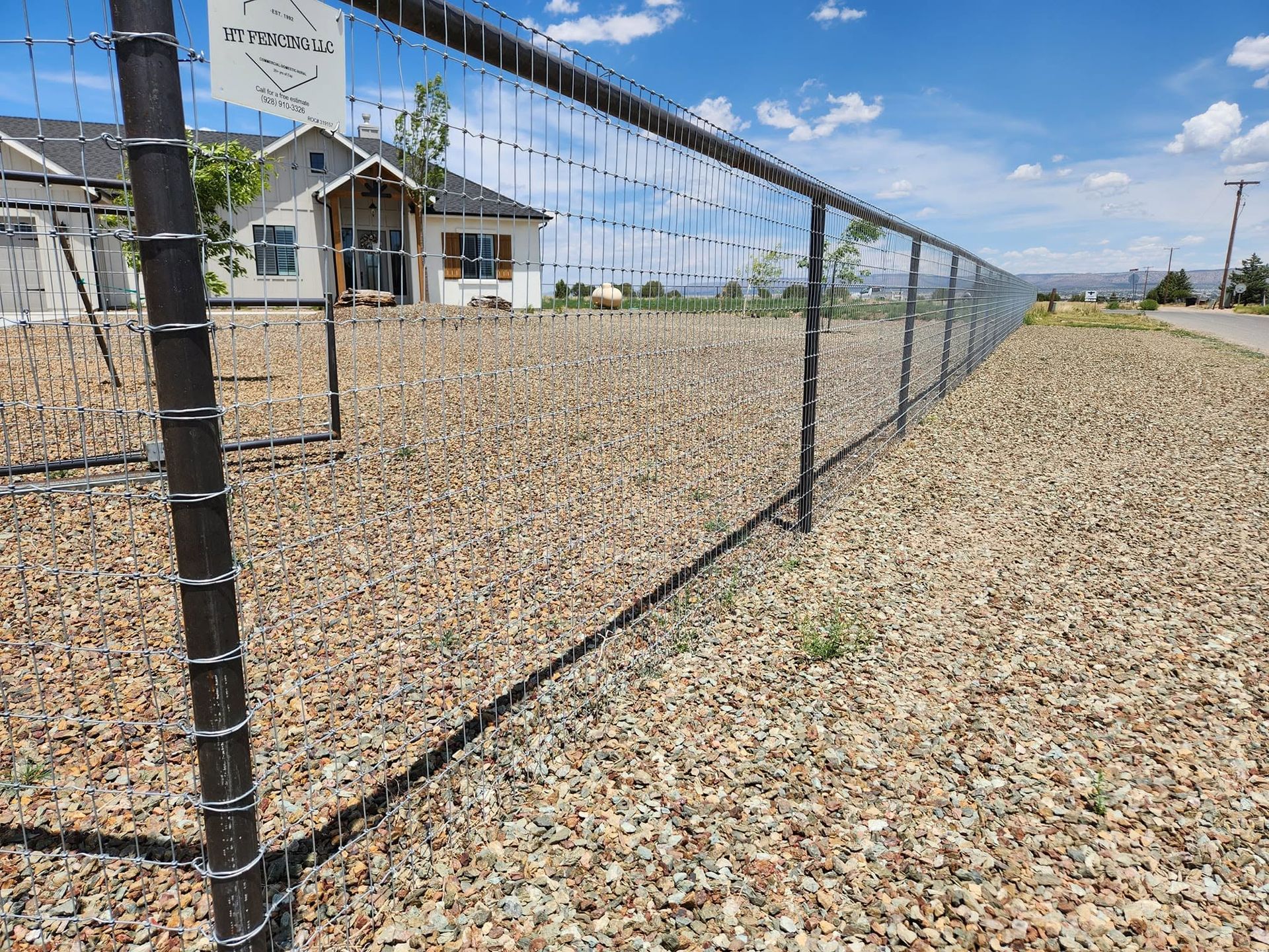 Chain-link fence beside a gravel lot with a house and blue sky in the background