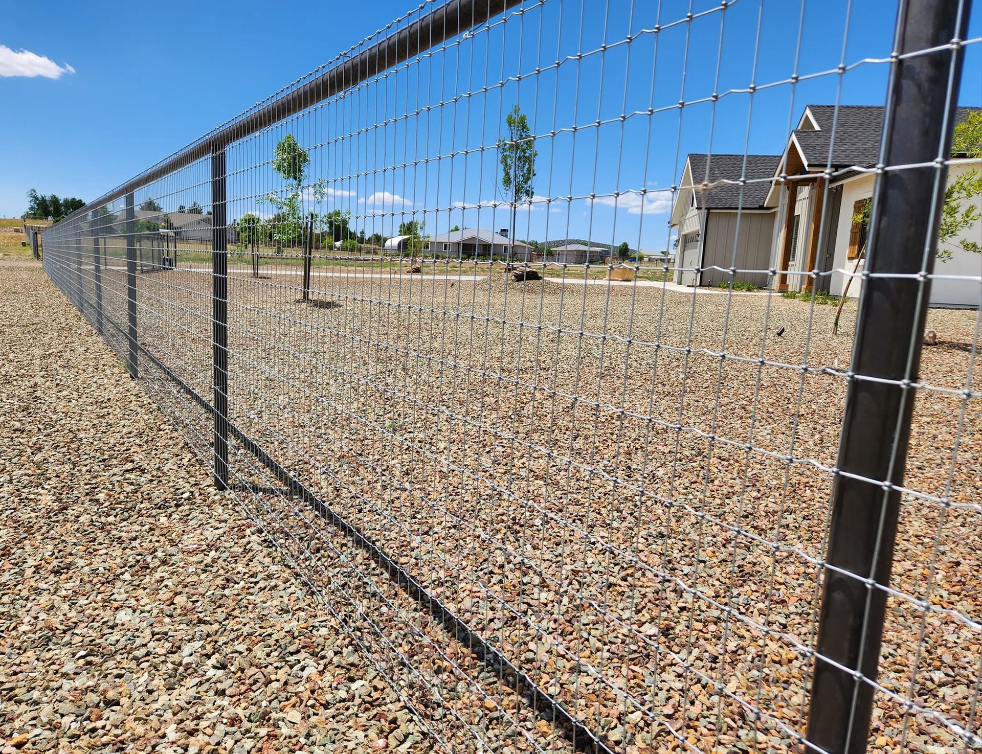 Wire mesh fence along a gravel yard beside a house under a clear blue sky.