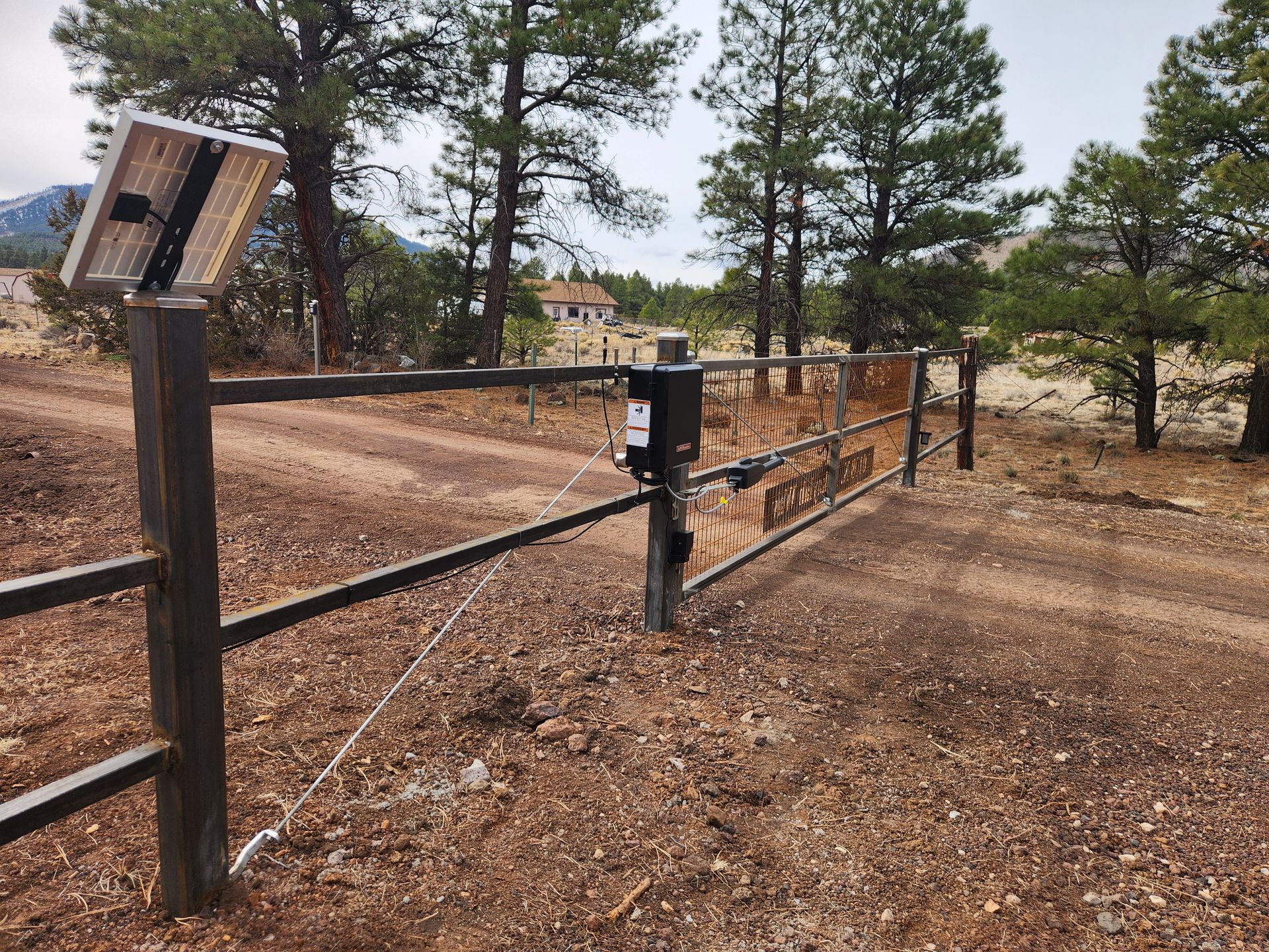 Rural dirt driveway blocked by a metal gate beside pine trees and a keypad post