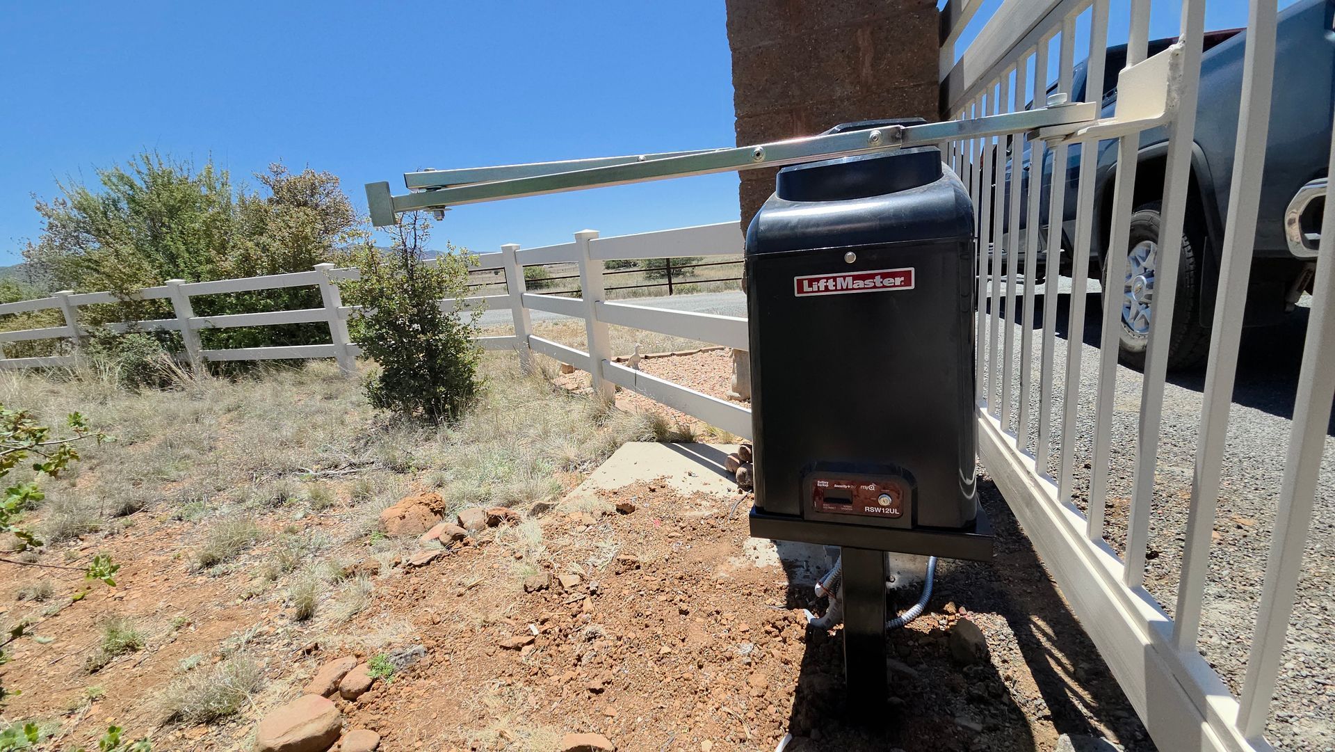 Black mailbox beside a white fence on a sunny dirt roadside, with a building and blue sky in the background