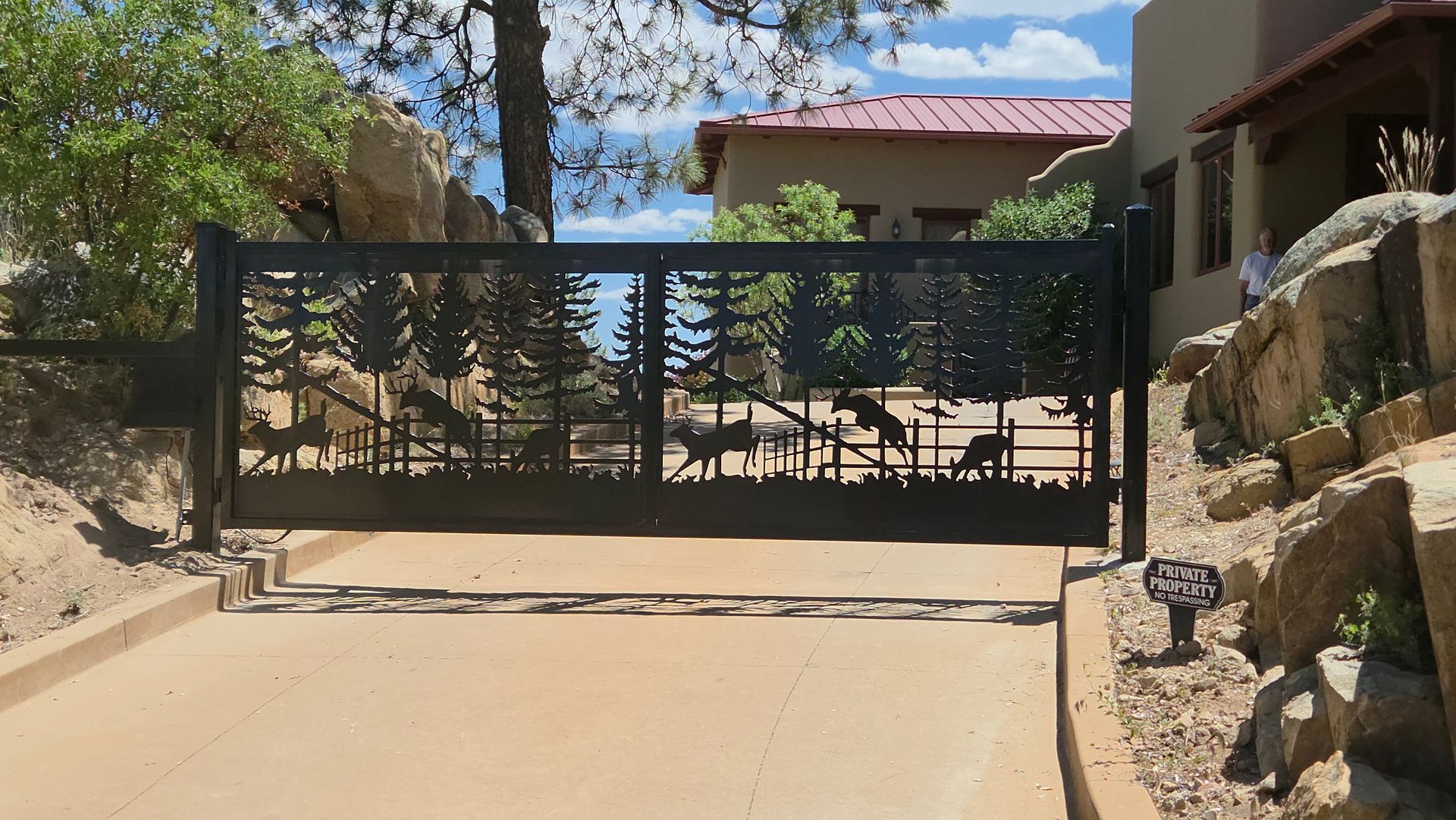 Black metal driveway gate at the end of a sloped sandy driveway, framed by rocks and trees.