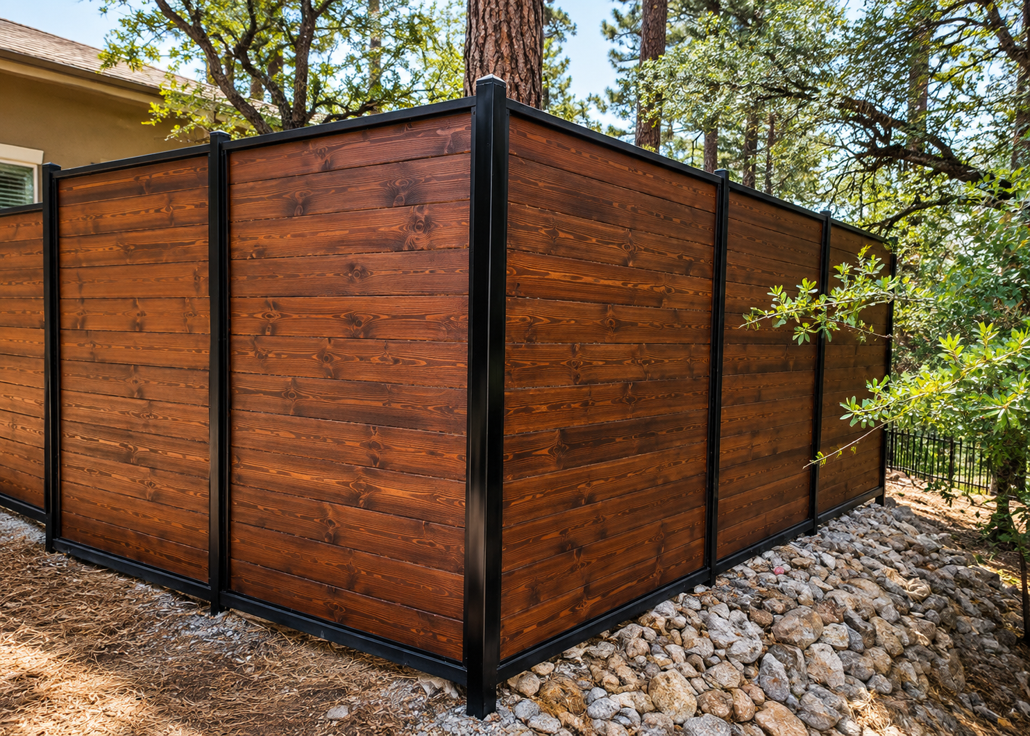 Wooden privacy fence with black metal posts beside a house and trees, set on a rock bed.