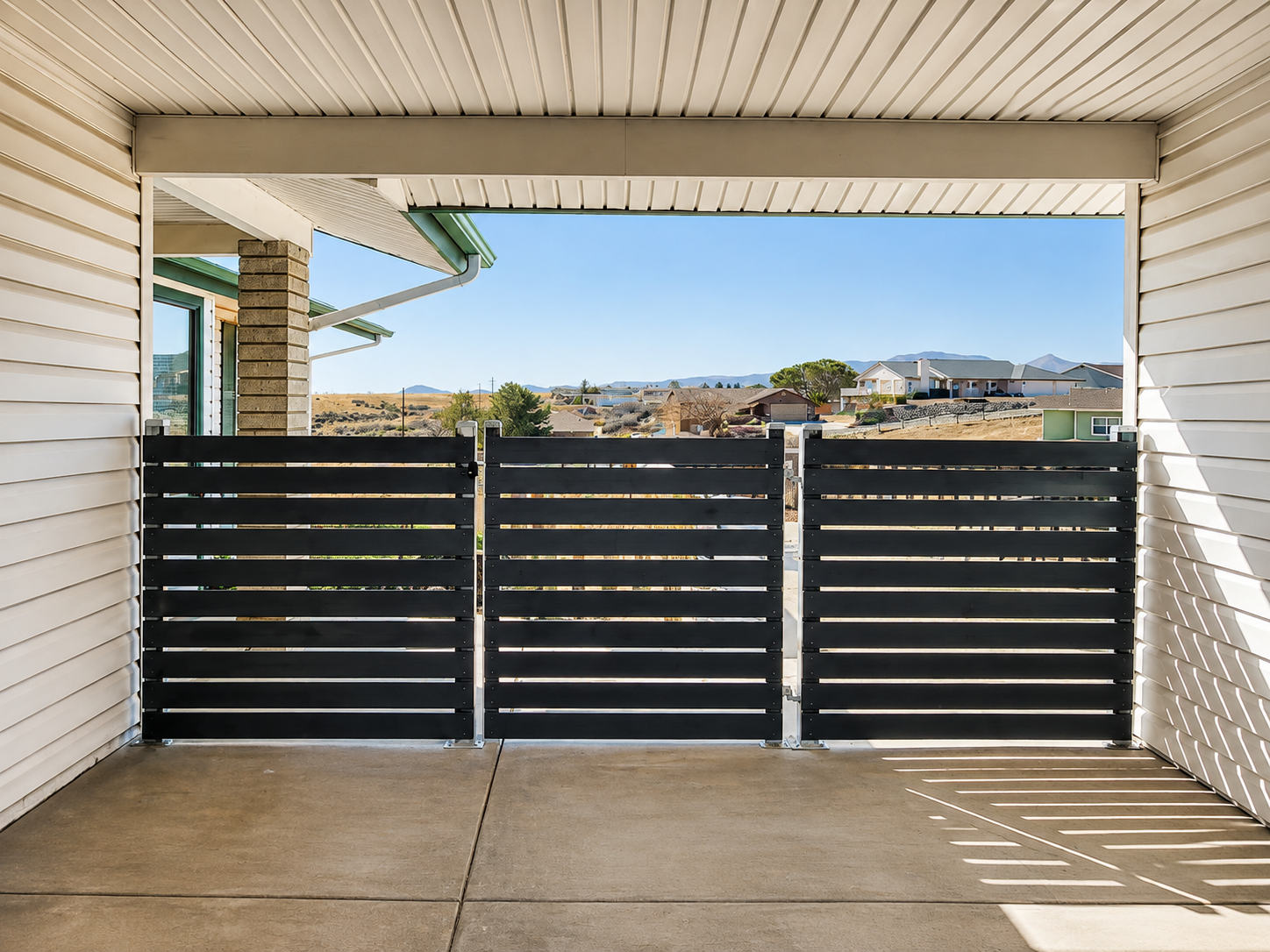 Covered patio with white siding, concrete floor, and black slatted gate overlooking a hillside view