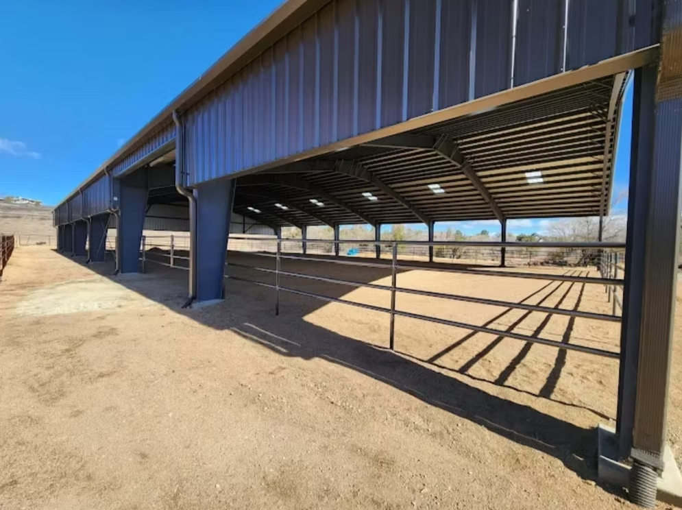 Blue metal open-sided barn on a dirt lot in a desert setting, with strong shadow lines from the roof