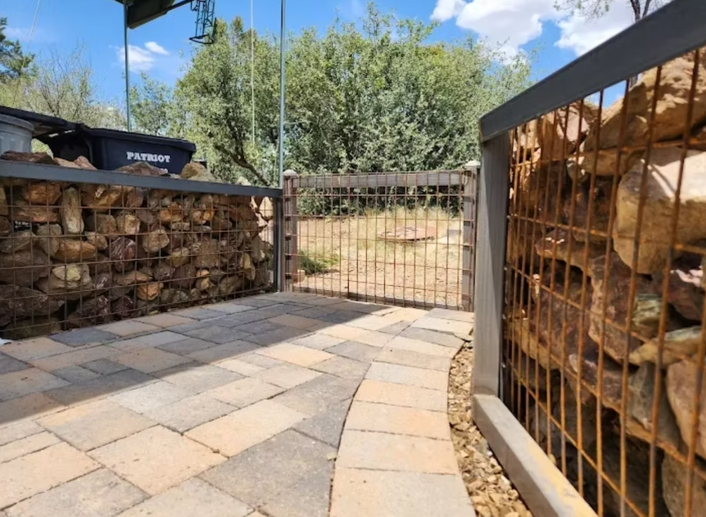 Stone patio with stacked firewood in a fenced yard under a sunny sky