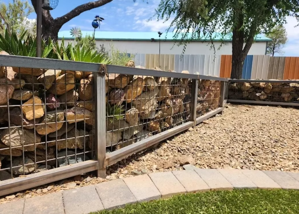 Woodpile stacked behind metal fencing along a gravel yard with trees and a building in the background