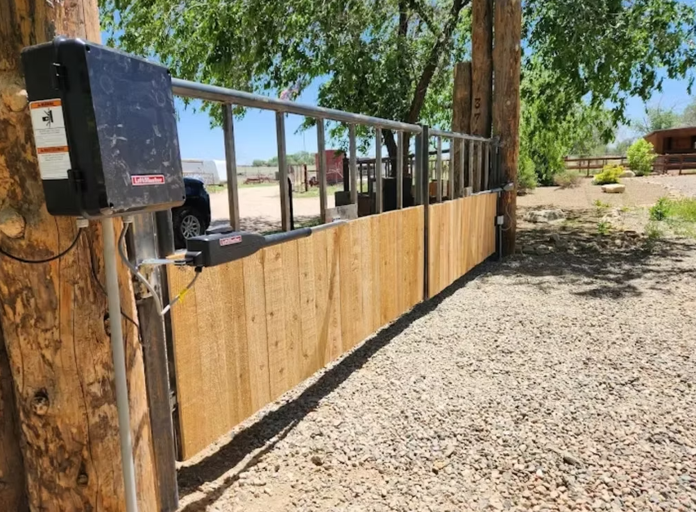 Wooden fence with black metal gate and mailbox beside a gravel driveway under trees