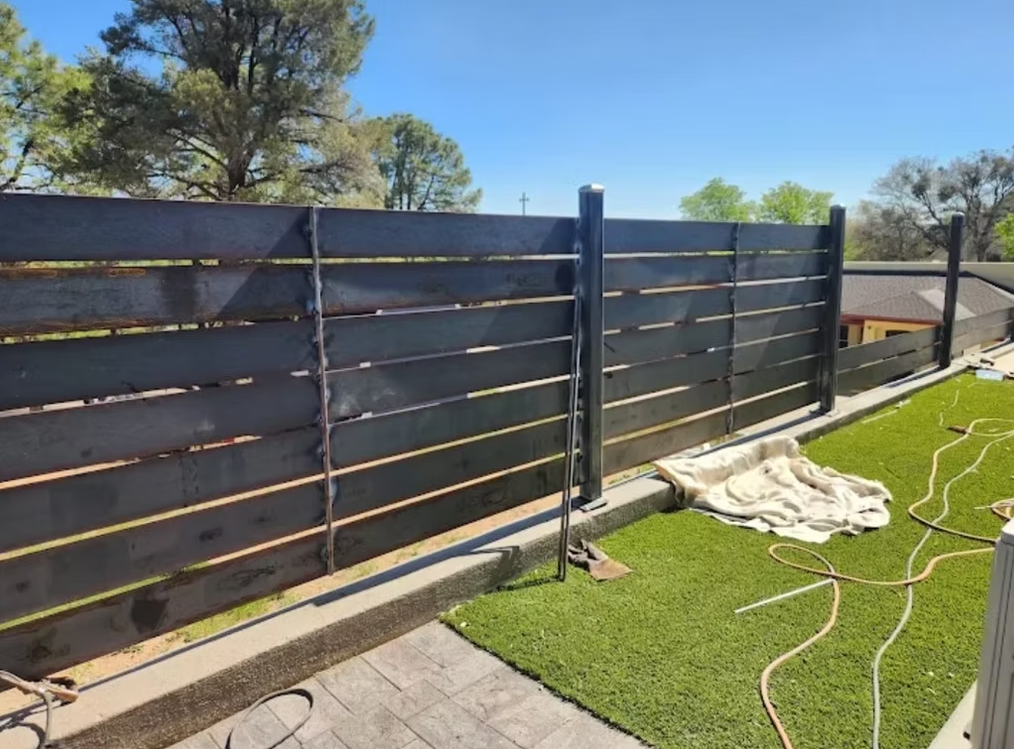 Black slatted fence along a patio beside artificial grass, with trees and a hose in the background