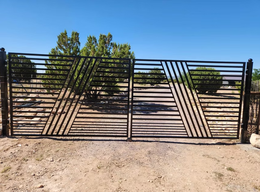 Black metal gate with diagonal slats across a dirt driveway under a clear blue sky.