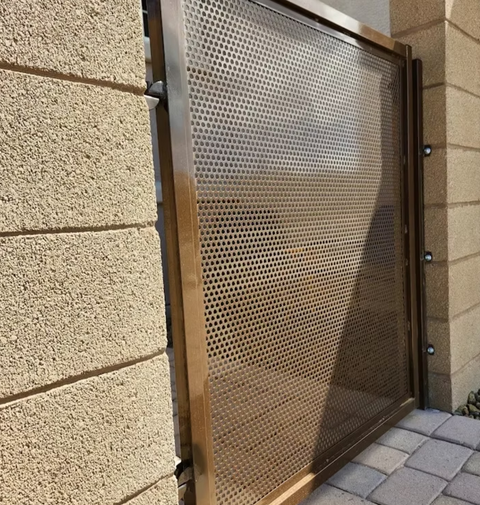 Perforated metal gate beside a tan stone wall on a paved walkway