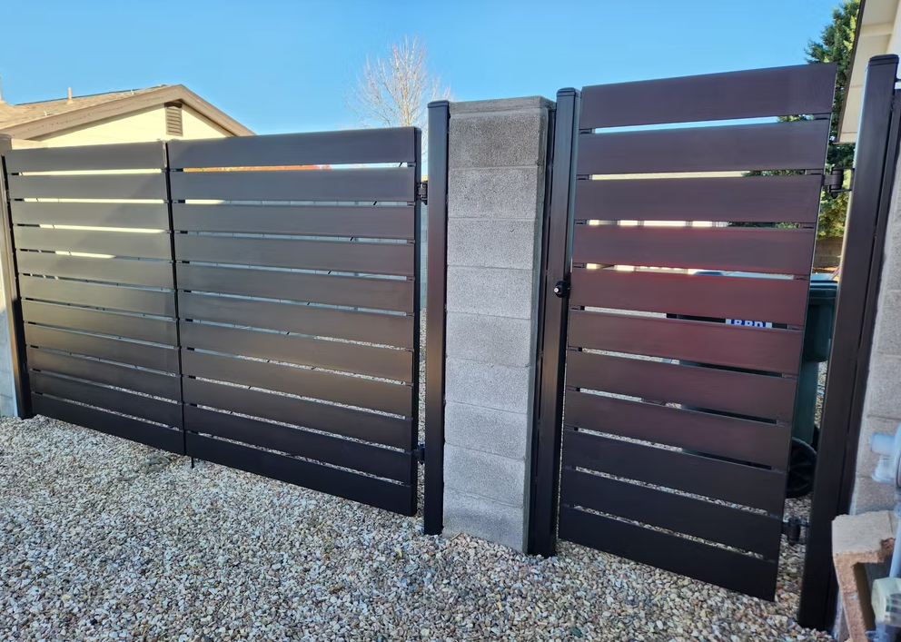 Modern gray slatted driveway gate with concrete pillar and gravel foreground, under a clear blue sky