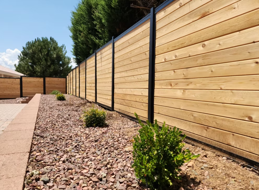 Wooden fence along a gravel yard with small shrubs and a concrete path under a clear blue sky